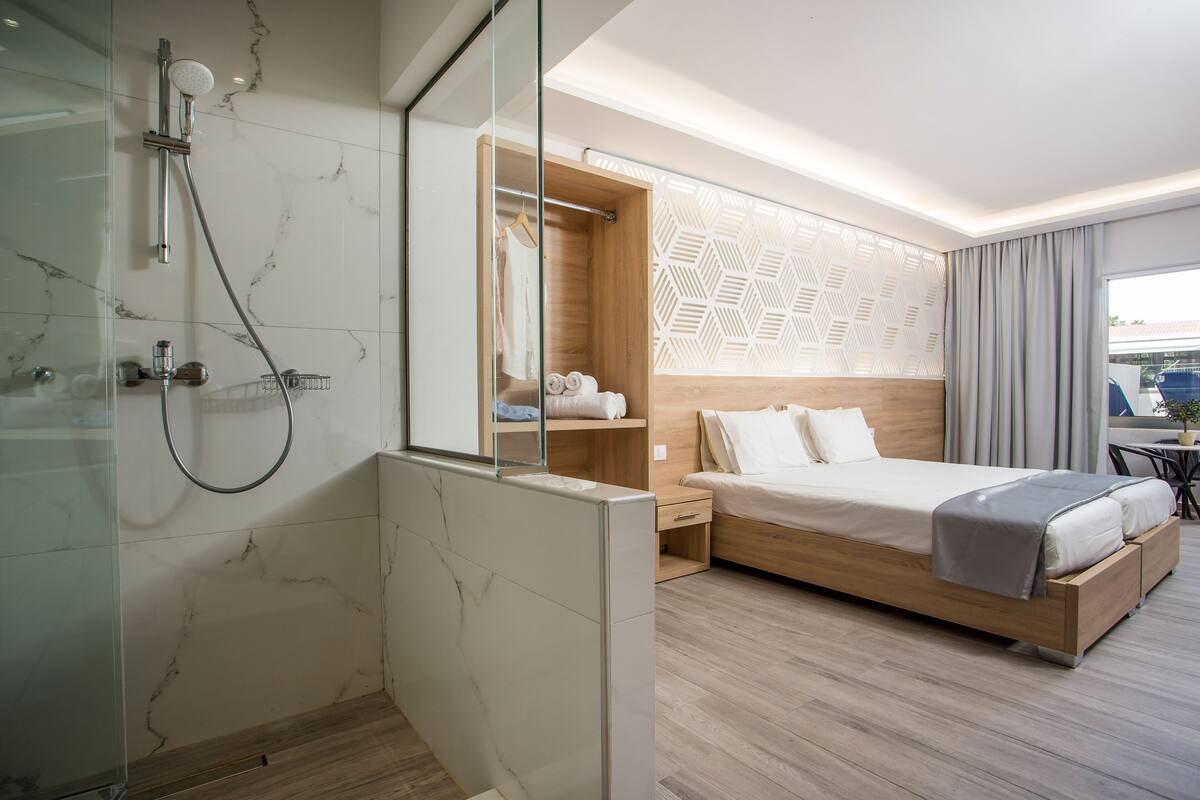 Una habitación de hotel moderna con ducha, cabecero de madera, cama con manta gris y un amplio ventanal con cortinas.