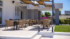 Un espace extérieur aménagé avec des chaises et des tables en osier, une pergola décorée de guirlandes lumineuses, et une vue sur la végétation et les bâtiments environnants.