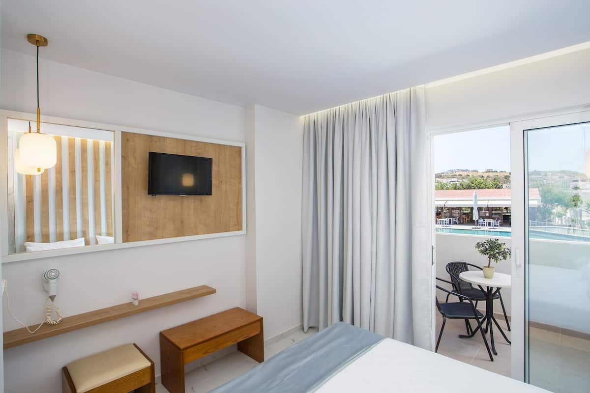 Una habitación de hotel moderna con una cama, un banco, una mesita con una planta, un televisor montado en un panel de madera y un balcón con vistas a la ciudad.