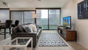 A modern living room with a leather sofa, a glass coffee table, a patterned rug, a TV on a wooden stand, and a large window with a view of greenery. - Condo magnifico in the heart of the Biltmore (Phoenix)