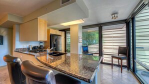 A modern kitchen with granite countertops, stainless steel appliances, and a large island. There are two bar stools and a dining area with a view through large sliding glass doors. - Condo magnifico in the heart of the Biltmore (Phoenix)