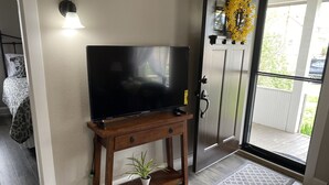 Een woonkamer met een flatscreen televisie op een houten standaard, een kamerplant en een deur die naar buiten leidt.