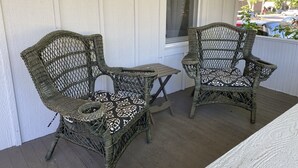 Twee rieten stoelen met gestreepte kussens op een houten terras, naast een witte muur en een raam.