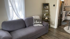 Een moderne woonkamer met een grijze bank, een klein houten plankje met decoratieve objecten, en een keuken die zichtbaar is door een open doorgang.