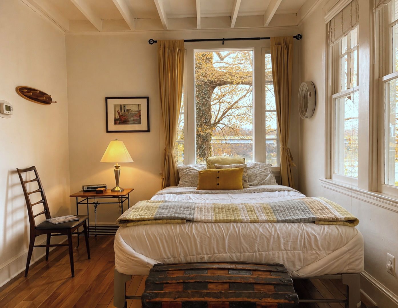 A bedroom with a bed, a chair, a bedside table with a lamp, a picture on the wall, and a window with curtains.