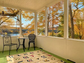 A room with a view of autumn trees and a bridge, a small table with a chair, and a patterned rug.