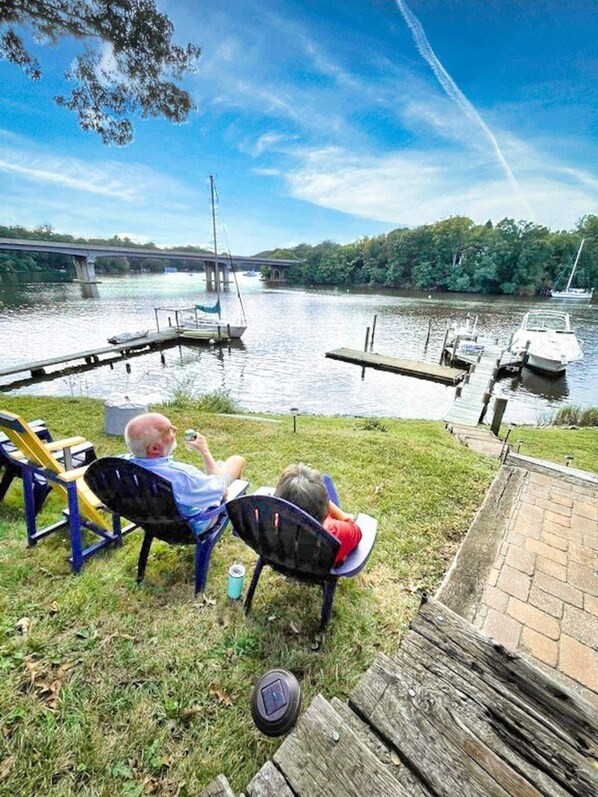 A riverside view with a bridge, boats, and a grassy area with chairs.