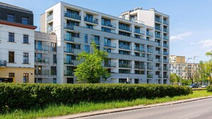 Un immeuble résidentiel à plusieurs étages, doté de balcons, avec un ciel dégagé et une haie le long de la rue.
