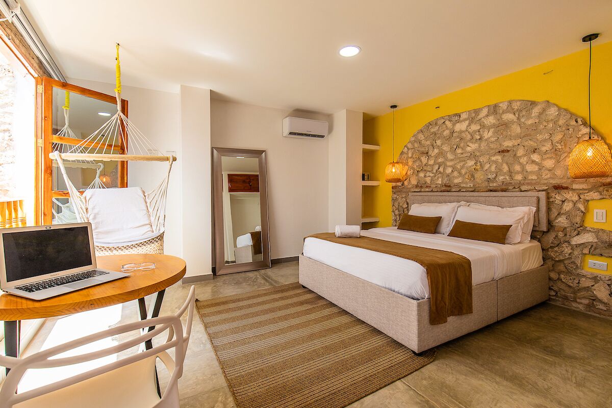 A bedroom with a bed, a desk with a laptop, a hammock, and a stone wall.