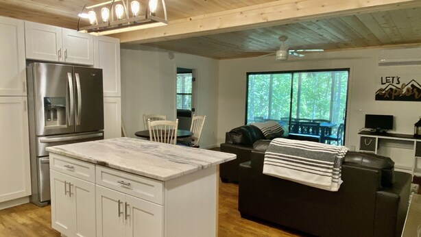 A modern kitchen with white cabinets, a stainless steel refrigerator, and a marble countertop. There is a dining area with a table and chairs, and a living room with a dark sofa and a window with a view of trees. - Shangri-La Cabin your mountain top Utopia (Blairsville)