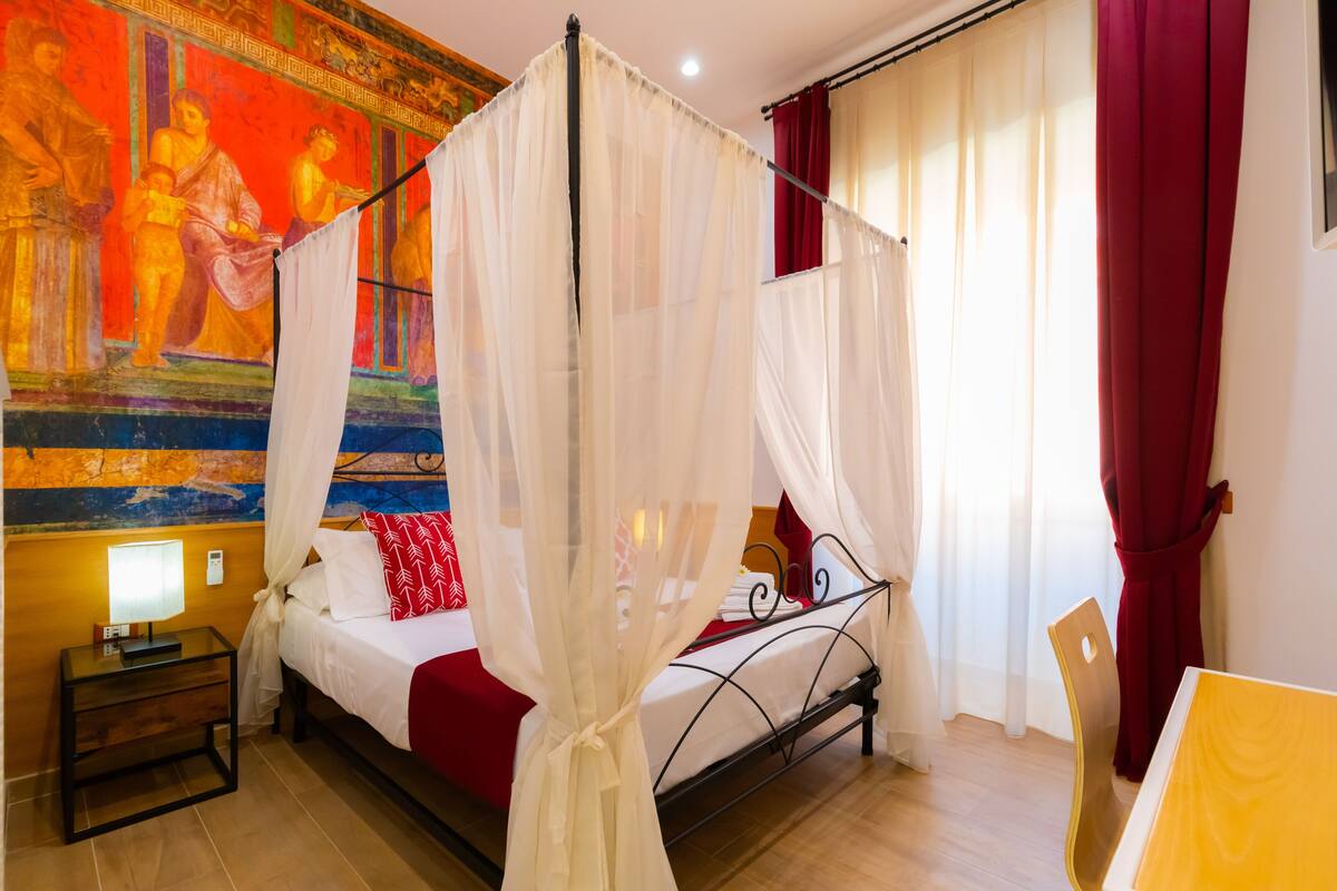 A room with a bed, a nightstand, a table, and a chair. There is a painting on the wall and a window with curtains.