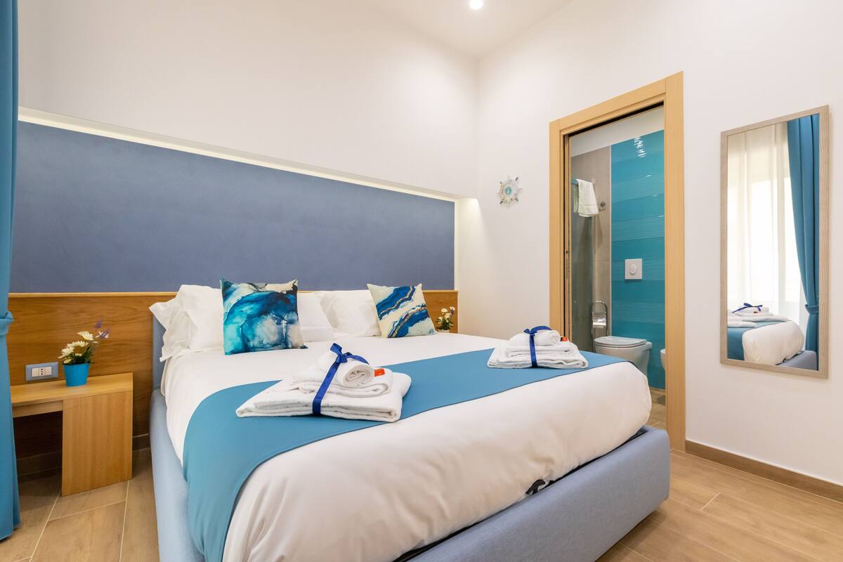 A hotel room with a large bed, a bedside table with a lamp, a mirror, and a bathroom visible through an open door.