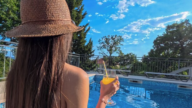 A person holding a drink by a pool with a clear sky and trees in the background. - Villa Mambo (Zupa dubrovacka)