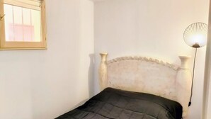 Ein kleines Zimmer mit einem Einzelbett, einem schlichten Kopfteil, einer Nachttischlampe und einem Fenster mit Jalousien.