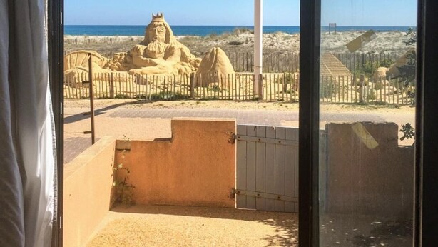 Blick durch ein Fenster auf eine Sandfläche mit einer Statue, einem Zaun und einer Straßenlaterne.