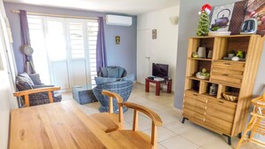 Un comedor con una mesa y sillas de madera, un sofá de mimbre, un televisor sobre un mueble bajo y un aparador de madera con objetos decorativos.