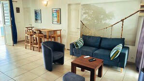 Una sala de estar con sofá, sillón, mesa de centro y una mecedora colgante. Hay una mesa de comedor con sillas y vistas al exterior a través de una ventana.