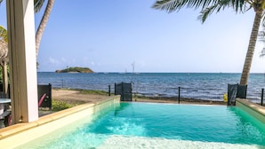 Piscina com água azul cristalina, cercada por uma grade e palmeiras, com vista para uma praia e oceano.