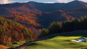 A golf course with a winding path, surrounded by autumn-colored trees and hills.