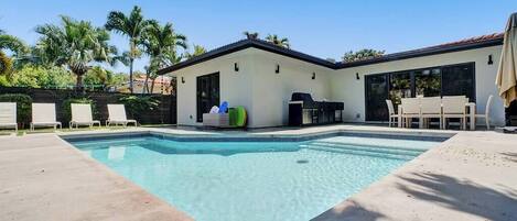 Piscina com espreguiçadeiras, área de churrasco e casa com grandes janelas.