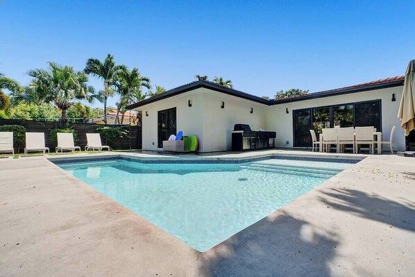 A swimming pool with lounge chairs, a barbecue area, and a house with large windows.