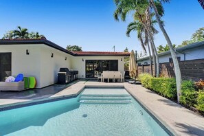 Une piscine avec des marches permettant d’y accéder, un patio avec des meubles d’extérieur et un espace barbecue couvert.