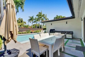 Une terrasse avec une table en verre et des chaises en osier, un barbecue et une piscine, avec des palmiers en arrière-plan.