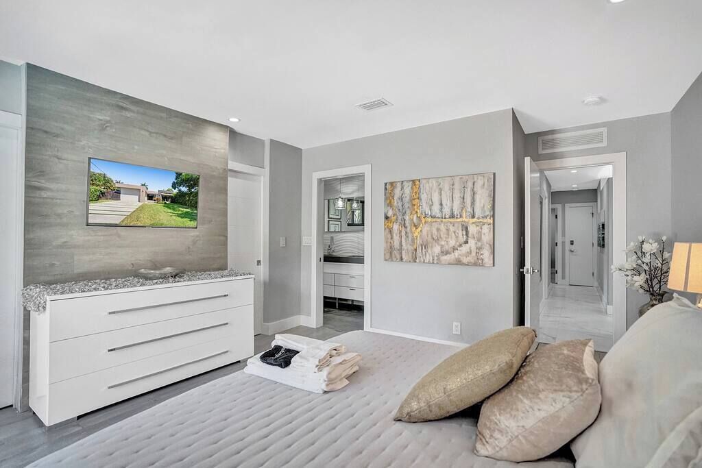 A modern bedroom with a large TV, a white dresser, a painting, and a view of the outside.