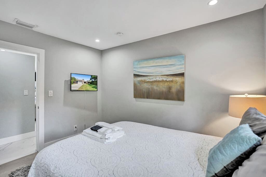 A bedroom with a bed, a TV mounted on the wall, and a painting above the bed.