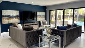 A modern living room with a large TV, comfortable seating, and a view of a swimming pool. - Welcome to Paradise! 1 mile to beach heated pool! (Hollywood)