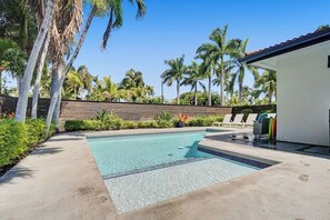 A swimming pool with a wooden fence, palm trees, and a white building with a red roof.