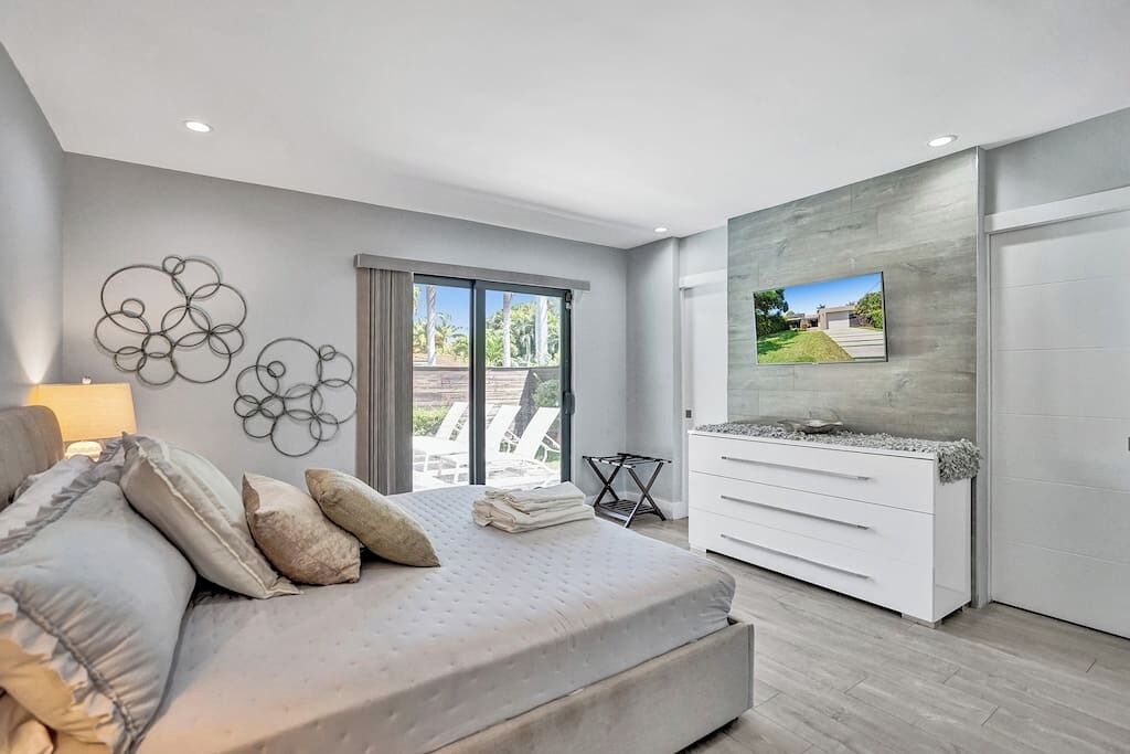 Une chambre moderne avec un lit, une télévision, une commode blanche et un balcon aménagé avec du mobilier d’extérieur.