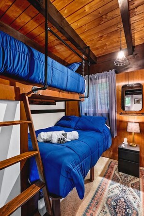 A bunk bed room with a wooden ceiling, a small bedside table with a lamp, and a patterned rug. - Chalet Getaway | Hot Tub, Sauna & Fire Pit (Bellaire)