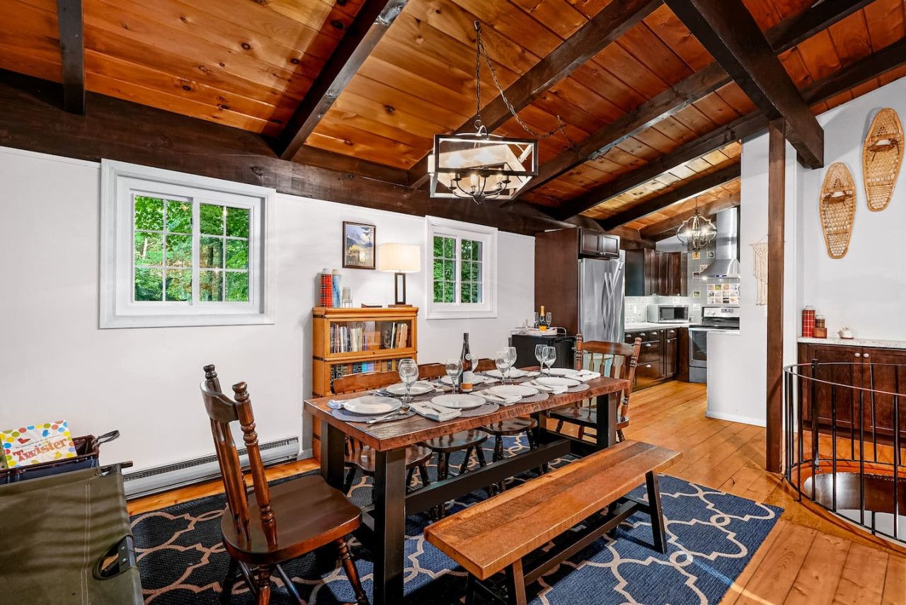 Un comedor con una mesa de madera dispuesta para seis personas, rodeada de sillas, un banco y una estantería.