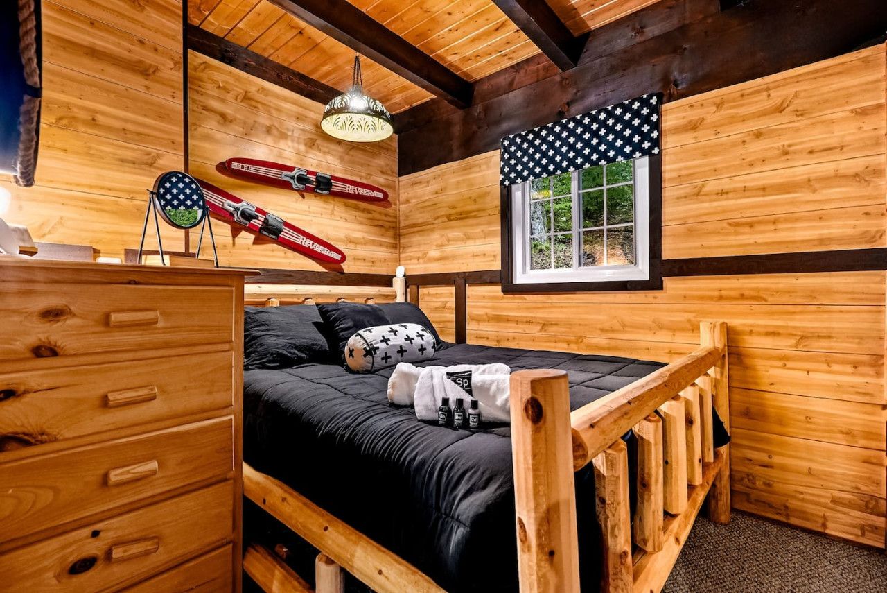 A wooden cabin room with a bed, a dresser, a window with a view, and skis hanging on the wall.