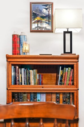 A wooden bookshelf with books and decorative items, a framed painting, and a table lamp. - Chalet Getaway | Hot Tub, Sauna & Fire Pit (Bellaire)