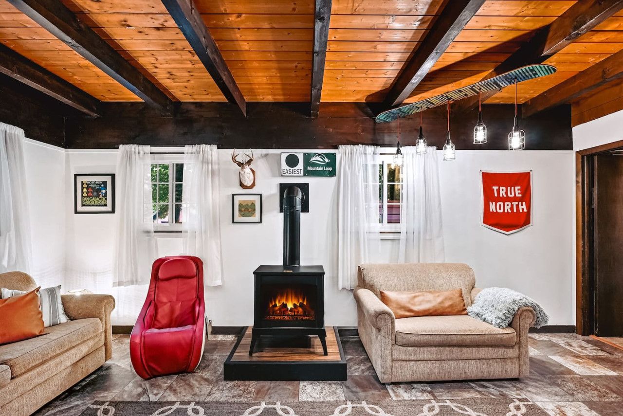 A cozy living room with a wood-burning stove, two sofas, a red chair, and a 'True North' sign.
