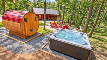 A wooden sauna structure, a hot tub, and outdoor seating with a red-roofed building in the background.