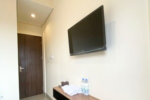 A hotel room with a mounted TV, a wooden bench with towels and bottled water, and a door in the background. - Reddoorz @ Arkana Seturan (Depok)