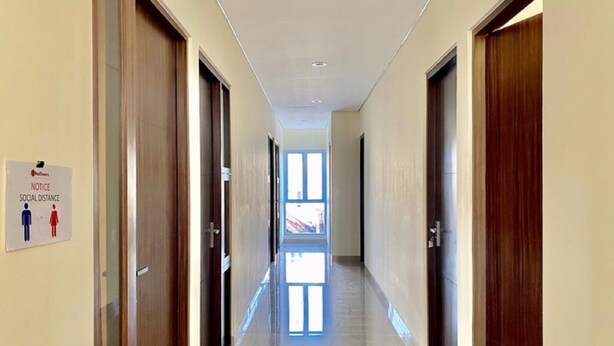 A hallway with doors on both sides, a sign indicating social distancing, and a window at the end. - Reddoorz @ Arkana Seturan (Depok)