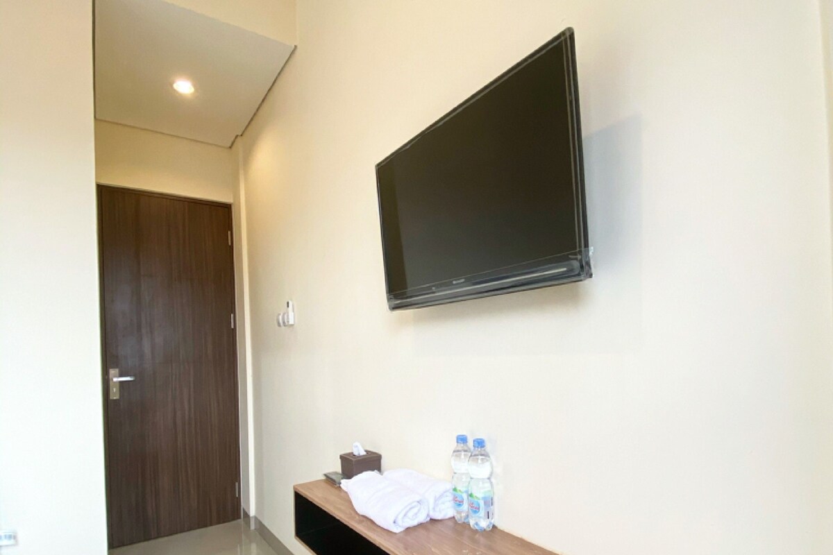 A hotel room with a mounted TV, a wooden bench with towels and bottled water, and a door in the background.