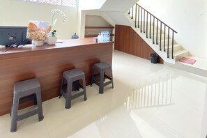 A reception area with a wooden counter, three stools, a desk with a computer, and a staircase. - Reddoorz @ Arkana Seturan (Depok)