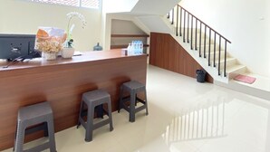 Zona de recepción con mostrador de madera, tres taburetes grises, una planta en maceta y una escalera que sube a un nivel superior.