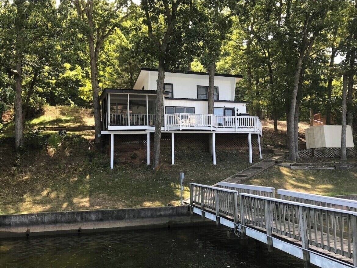 A modern two-story house with a balcony, surrounded by trees and a dock over water.