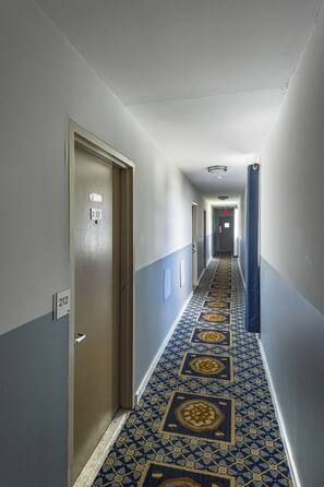 Un couloir d’hôtel avec un tapis à motifs, des portes numérotées 212 et un rideau bleu sur la droite.