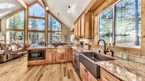 A spacious kitchen with wooden cabinets, a large sink, and a microwave. There are large windows offering views of trees.