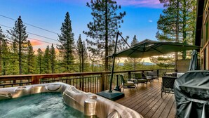 A hot tub on a wooden deck with outdoor furniture and a view of trees and mountains.