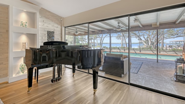 A spacious room with a grand piano, large windows, and a view of a pool and ocean. - NOW ACCEPTING ONE YEAR LEASES ON OUR WATERFRONT HOME! (Merritt Island)