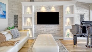 A modern living room with a flat-screen TV, a white sofa, a glass-top coffee table, and a grand piano. - NOW ACCEPTING ONE YEAR LEASES ON OUR WATERFRONT HOME! (Merritt Island)