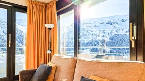 Ein gemütliches Zimmer mit einer Couch, einer Lampe und großen Fenstern mit Blick auf einen verschneiten Berg.